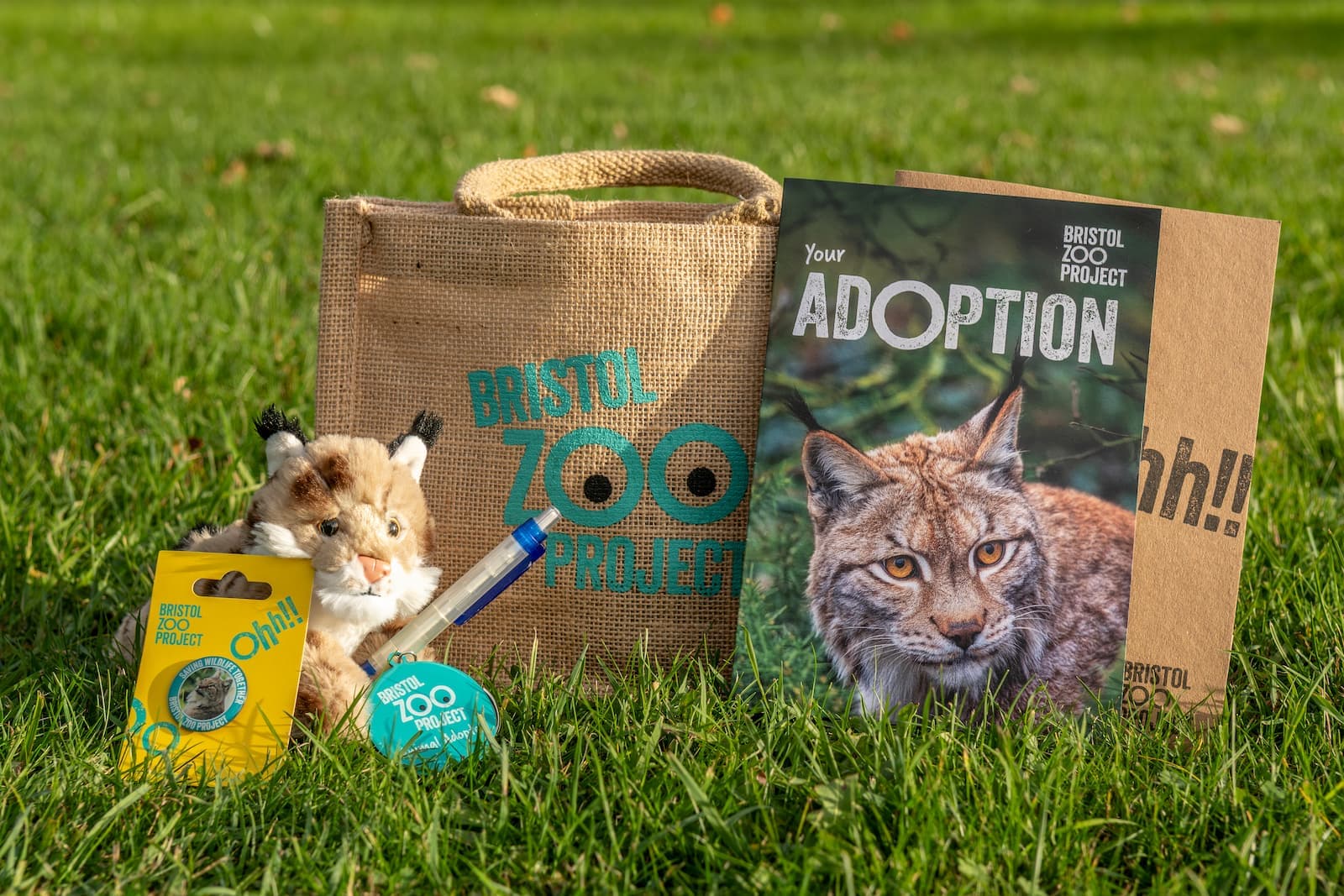 Bristol Zoo Project adoption kit on grass, includes a lynx photo, plush toy, jute bag, and informational materials.