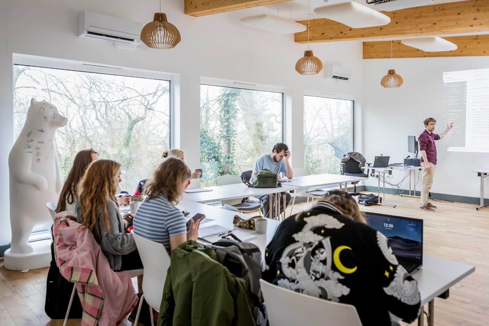Classroom with students sitting at tables, using laptops and notebooks. A teacher stands near a projector screen. Large windows show trees outside.